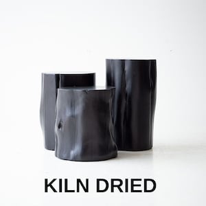 May include: Three black wooden stools, each with a different height, are arranged on a white surface. The stools are made from natural wood and have a rough, organic texture. The text "KILN DRIED" is printed below the stools.