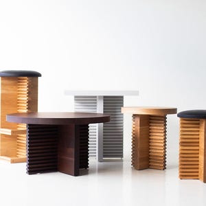 May include: A collection of modern wooden furniture pieces, including a round coffee table, a rectangular side table, and two stools with black padded seats. The tables and stools have a slatted design.