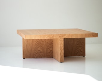 Puede incluir: Una mesa de centro cuadrada de madera con un diseño de base abierto único. La mesa está hecha de madera marrón claro con un patrón de veta natural.