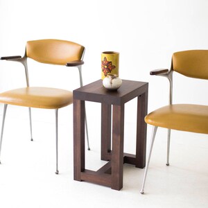 Puede incluir: Una mesa auxiliar de madera marr&oacute;n oscuro con una tapa cuadrada y un dise&ntilde;o de marco simple y abierto. Dos sillas de color amarillo mostaza con patas plateadas se colocan a cada lado de la mesa. Un jarr&oacute;n de cer&aacute;mica amarillo con un dise&ntilde;o de flor roja y un peque&ntilde;o cuenco de cer&aacute;mica blanco est&aacute;n sobre la mesa.