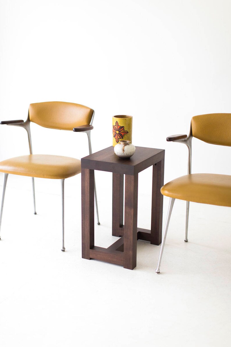 Puede incluir: Una mesa auxiliar de madera marr&oacute;n oscuro con una tapa cuadrada y un dise&ntilde;o de marco simple y abierto. Dos sillas de color amarillo mostaza con patas plateadas se colocan a cada lado de la mesa. Un jarr&oacute;n de cer&aacute;mica amarillo con un dise&ntilde;o de flor roja y un peque&ntilde;o cuenco de cer&aacute;mica blanco est&aacute;n sobre la mesa.