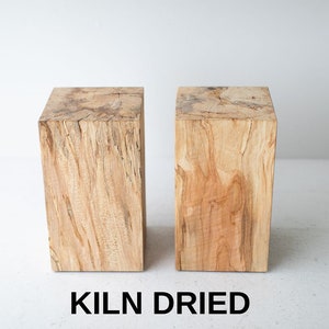 May include: Two natural wood blocks, each with a unique grain pattern, are shown on a white surface. The blocks are square and have a rough, unfinished texture. The text "KILN DRIED" is printed below the blocks.