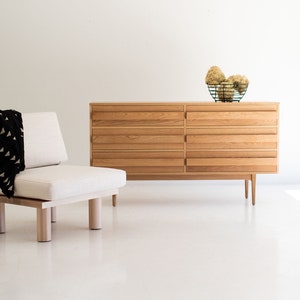 May include: A light wood dresser with six drawers and a simple design. The dresser is sitting on a white floor and has a small, white ottoman in front of it.