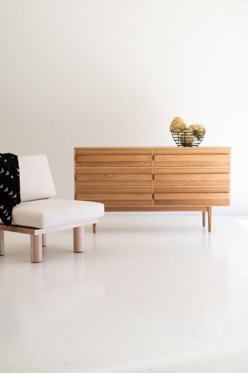 May include: A light wood dresser with six drawers and a simple design. The dresser is sitting on a white floor and has a small, white ottoman in front of it.