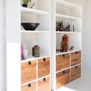 May include: White bookshelf with wooden drawers and shelves. The shelves are decorated with various items, including a black bowl, a ceramic vase, and a glass bottle. The drawers have black leather pulls.