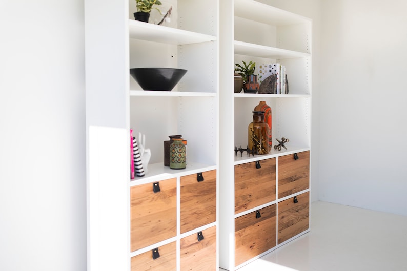 May include: White bookshelf with wooden drawers and shelves. The shelves are decorated with various items, including a black bowl, a ceramic vase, and a glass bottle. The drawers have black leather pulls.