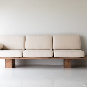 Pode incluir: Um sof&aacute; de madeira castanho claro com um almofada de tecido branco e uma almofada a condizer. O sof&aacute; tem um perfil baixo e um design simples.