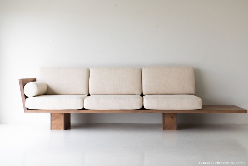 Pode incluir: Um sof&aacute; de madeira castanho claro com um almofada de tecido branco e uma almofada a condizer. O sof&aacute; tem um perfil baixo e um design simples.