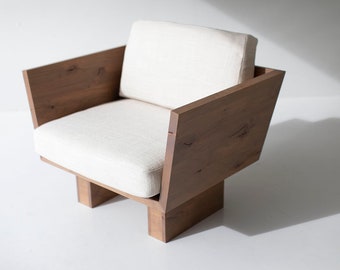 May include: A modern armchair with a solid wood frame and a cream-colored cushion. The chair features a geometric design with angled armrests and a square seat. The wood has a natural grain and a warm, brown tone.
