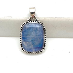 May include: A rectangular pendant with a blue gemstone set in a silver-colored metal frame. The gemstone has a cloudy appearance with shades of blue. The frame is adorned with small, round details and a loop for a chain.