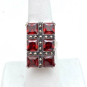 May include: A silver ring with a red gemstone set in a square shape. The gemstone is surrounded by a border of smaller silver stones.