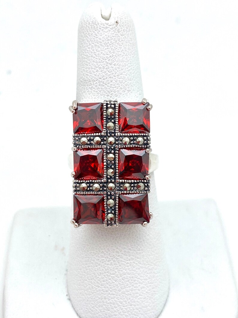 May include: A silver ring with a red gemstone set in a square shape. The gemstone is surrounded by a border of smaller silver stones.