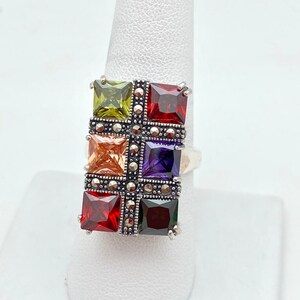 May include: A silver ring with a rectangular design featuring six square-shaped gemstones in various colors, including green, orange, purple, and red. The ring is set with marcasite stones.