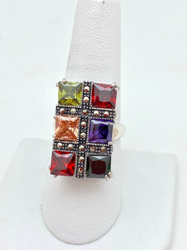May include: A silver ring with a rectangular design featuring six square-shaped gemstones in various colors, including green, orange, purple, and red. The ring is set with marcasite stones.