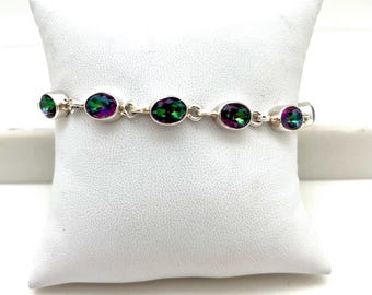 Peut inclure: Un bracelet en argent orné de pierres précieuses ovales et irisées aux tons verts, violets et bleus. Les pierres sont serties dans un design en chaîne, créant un bijou délicat et élégant. Le bracelet est présenté sur un coussin blanc.