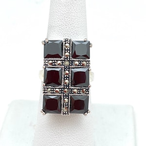 May include: A silver ring with a black gemstone set in a square shape. The gemstone is surrounded by smaller silver stones.
