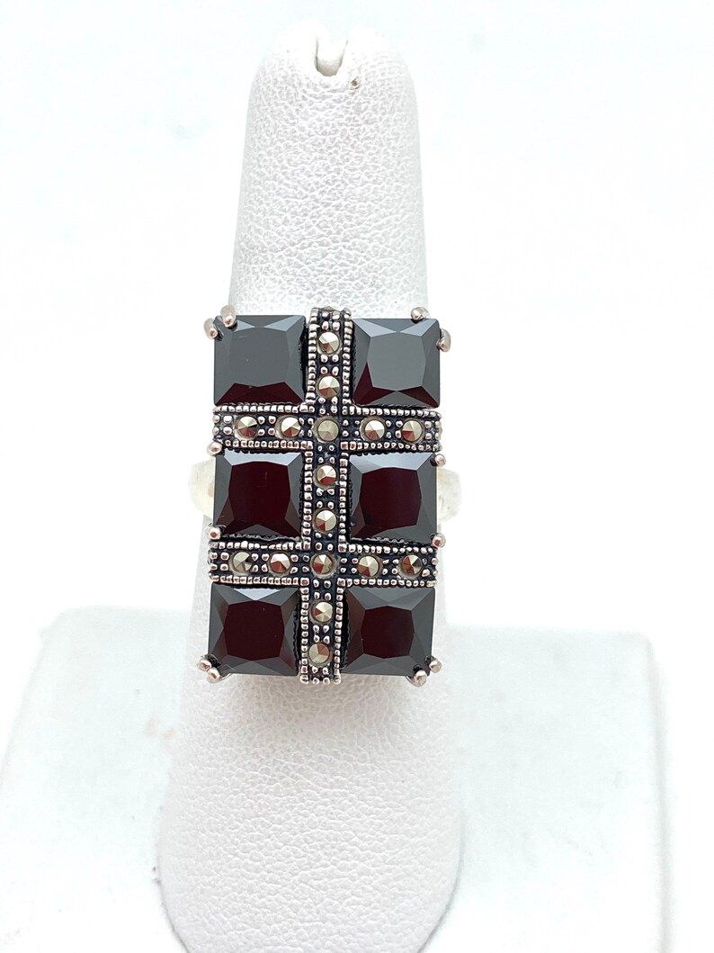 May include: A silver ring with a black gemstone set in a square shape. The gemstone is surrounded by smaller silver stones.