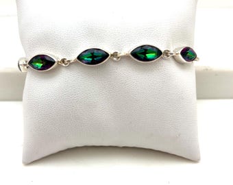 Peut inclure: Un bracelet en argent orné de pierres précieuses ovales et irisées, aux teintes vertes, violettes et bleues. Les pierres sont reliées par de petits maillons en argent. Le bracelet est présenté sur une surface blanche texturée.
