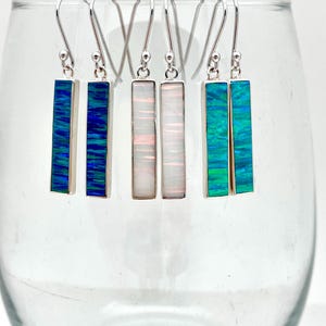 May include: Five pairs of silver dangle earrings with rectangular opal stones. The opals are in shades of blue, green, and white.