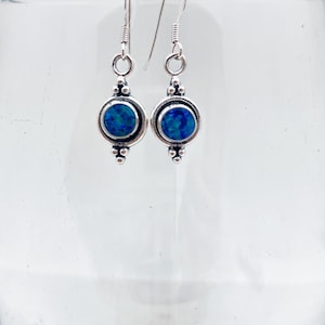 May include: A pair of silver-toned dangle earrings. Each earring features a round, bezel-set, blue gemstone with a speckled appearance. The earrings have decorative details and a hook closure.