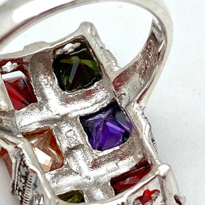 May include: A silver ring with a rectangular design featuring five square-shaped gemstones. The gemstones are red, green, purple, and two clear stones. The ring is decorated with small black stones.