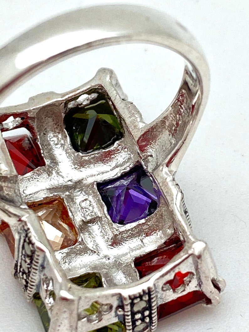 May include: A silver ring with a rectangular design featuring five square-shaped gemstones. The gemstones are red, green, purple, and two clear stones. The ring is decorated with small black stones.