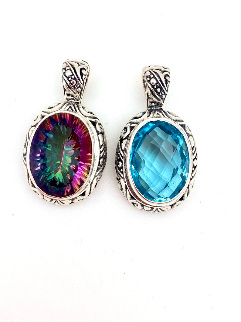 May include: Two silver pendants with oval-shaped gemstones. The pendant on the left has a rainbow mystic topaz gemstone. The pendant on the right has a light blue topaz gemstone.