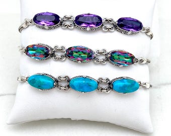 Peut inclure: Trois bracelets en argent avec des pierres précieuses ovales. Le bracelet supérieur présente des améthystes violettes, le milieu des pierres irisées et le bas des turquoises. Chaque bracelet est présenté sur un coussin blanc.