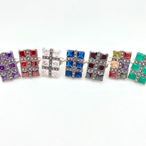 May include: Six silver rings with a square design. Each ring features a different colored gemstone in the center, surrounded by smaller white gemstones. The gemstones are purple, red, white, blue, black, green, and yellow.