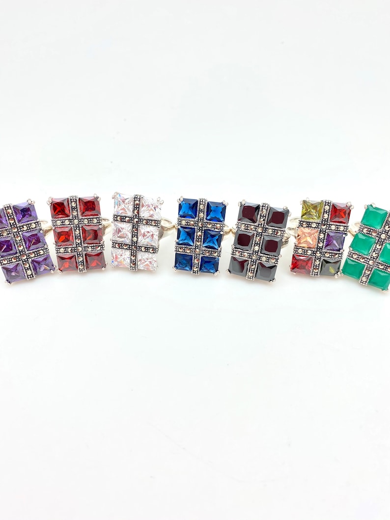 May include: Six silver rings with a square design. Each ring features a different colored gemstone in the center, surrounded by smaller white gemstones. The gemstones are purple, red, white, blue, black, green, and yellow.