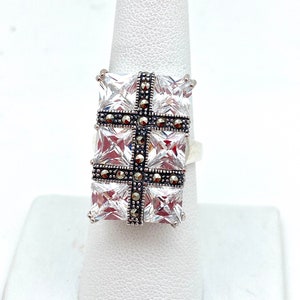 May include: A silver ring with a large square-shaped clear stone set in the center. The stone is surrounded by smaller black stones in a cross pattern.