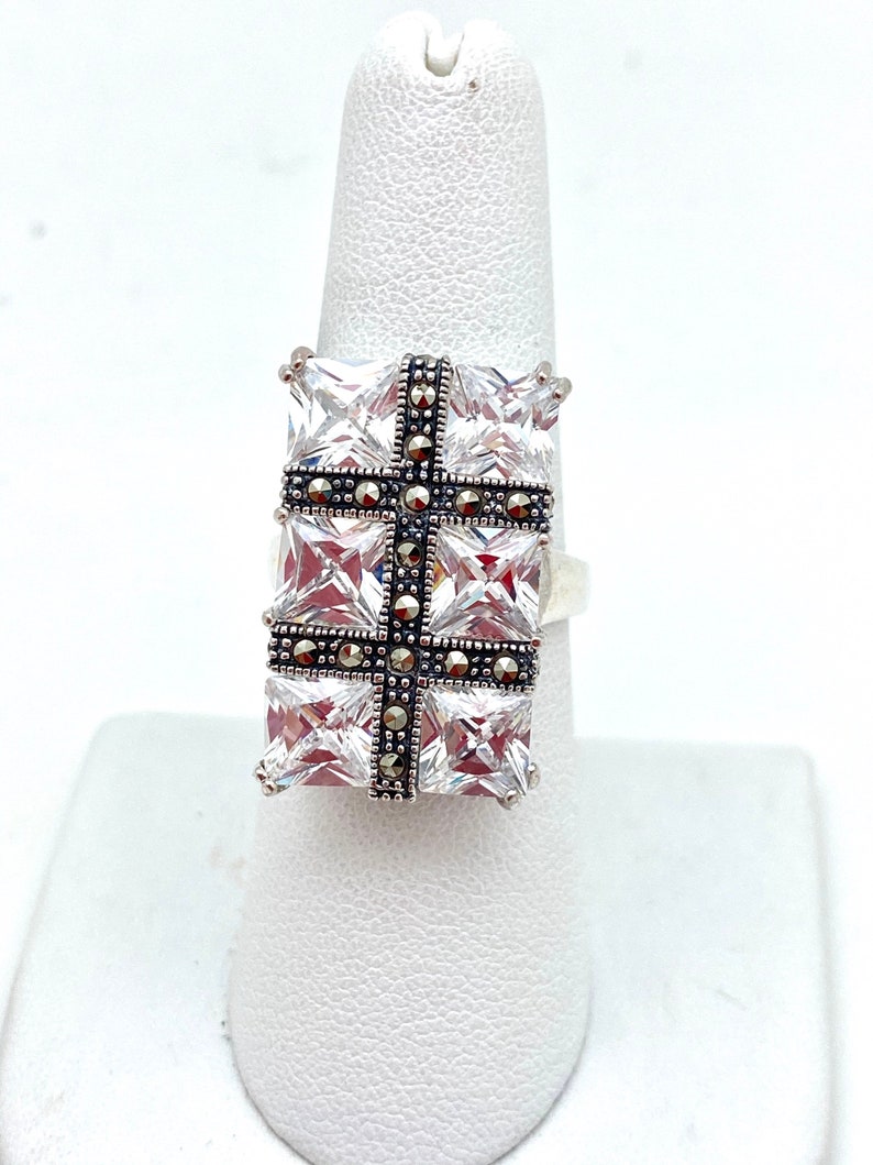 May include: A silver ring with a large square-shaped clear stone set in the center. The stone is surrounded by smaller black stones in a cross pattern.