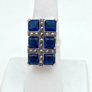 May include: A silver ring with a blue stone cross design. The cross is made of four square blue stones surrounded by smaller silver stones.