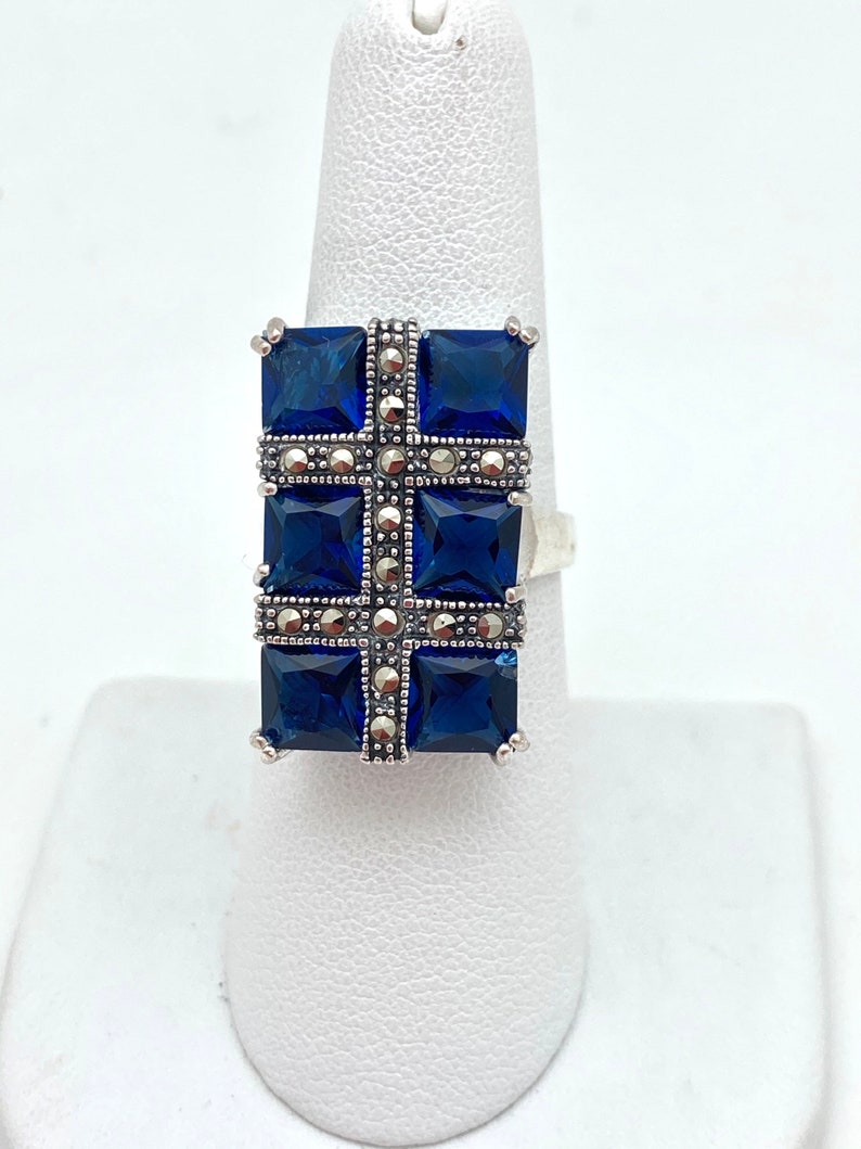 May include: A silver ring with a blue stone cross design. The cross is made of four square blue stones surrounded by smaller silver stones.