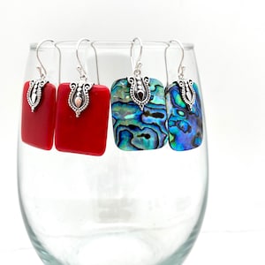 May include: Three pairs of earrings with silver hooks and rectangular shaped stones. One pair has red stones, one pair has blue and green abalone shell stones, and one pair has blue abalone shell stones.