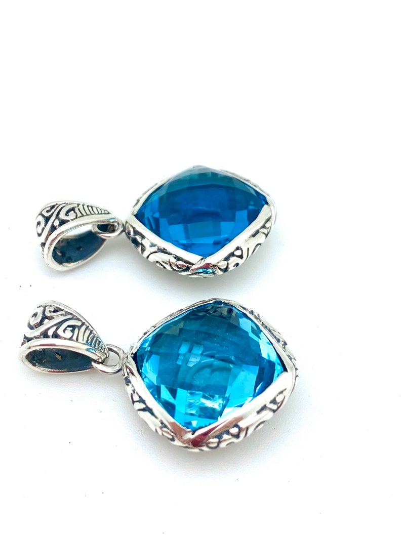 May include: Two silver pendants with a blue gemstone in a square shape. The pendants have an intricate silver design around the gemstone.