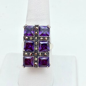 May include: A silver ring with a square-shaped center stone set with multiple smaller purple stones. The center stone is surrounded by a border of smaller, gray stones.