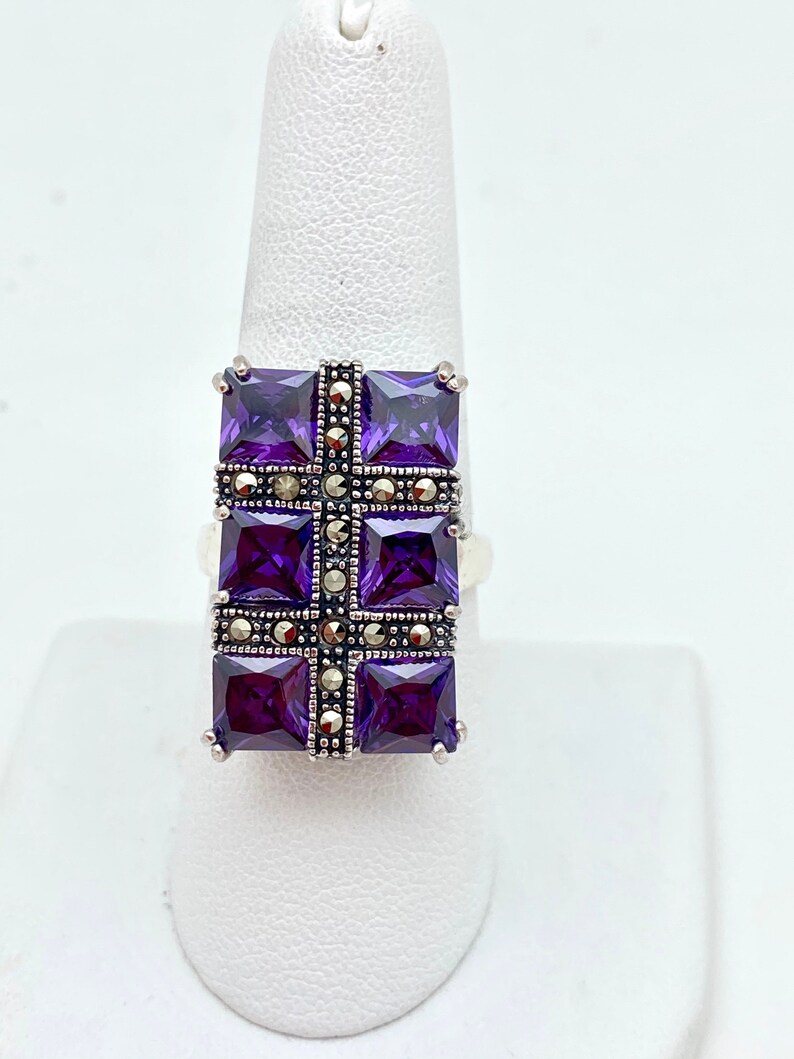 May include: A silver ring with a square-shaped center stone set with multiple smaller purple stones. The center stone is surrounded by a border of smaller, gray stones.