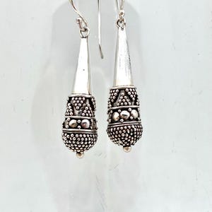 May include: A pair of silver dangle earrings with a textured, beaded design. The earrings are teardrop shaped and have a silver hook closure.