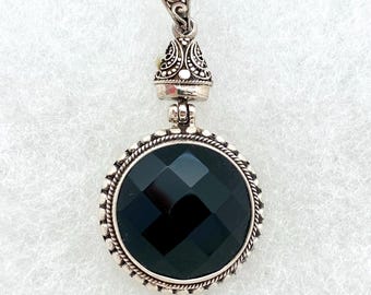 May include: A silver pendant with a black gemstone set in a round bezel. The pendant has a detailed filigree design and a bail for attaching to a chain.
