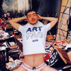 May include: A person wearing a white t-shirt with the text "ART FAG GUTS" in blue lettering. The person is sitting in a cluttered room with paint supplies and other items.