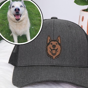 以下が含まれることがあります： 笑顔のハスキー犬の顔が描かれた茶色の革パッチが付いた、チャコールグレーのトラッカーハット。帽子はメッシュの背面と湾曲したつばが付いています。ハスキー犬の円形の画像が背景にあります。