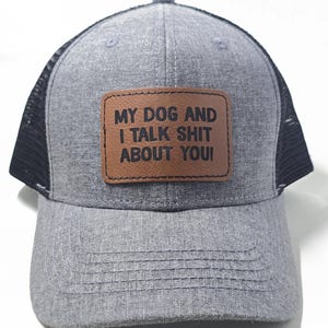 May include: Gray trucker hat with a black mesh back and a curved brim. The hat features a brown leather patch with the text "MY DOG AND I TALK SHIT ABOUT YOU!" in white letters. The hat is a casual accessory.
