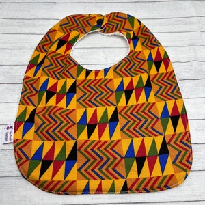 May include: A baby bib with a vibrant geometric pattern in yellow, red, blue, green, and black. The bib has a rounded shape with a neck opening and a snap closure. The bib is made of fabric and has a white backing.