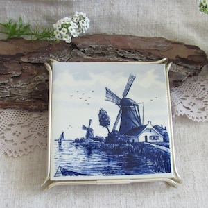 Può includere: Piastrella quadrata in ceramica con una scena di mulino a vento blu e bianca, incorniciata in una cornice di metallo argentato. La piastrella raffigura un paesaggio in riva al mare con mulini a vento, una casa e una barca a vela. Fiori bianchi e corteccia sullo sfondo.