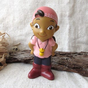 Può includere: Una statuina di un personaggio dei cartoni animati con una camicia rosa, pantaloni blu scuro e stivali marroni. Il personaggio ha un copricapo rosa e un ciondolo a fischietto giallo. La statuina è in piedi su un pezzo di legno.