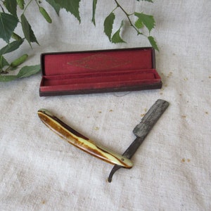 May include: An antique straight razor with a cream and brown handle, open and ready for use. The blade is silver and shows signs of age. A small, open, brown case with a red interior sits nearby. The razor is on a white cloth.