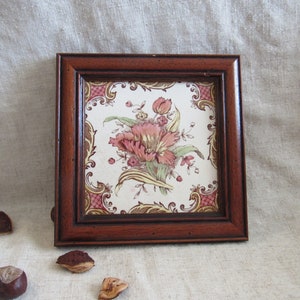 May include: Square wooden framed tile with a floral design. The tile features a central arrangement of pink and green flowers on a white background, framed by a dark brown wooden frame. Decorative accents in pink and gold surround the floral image.