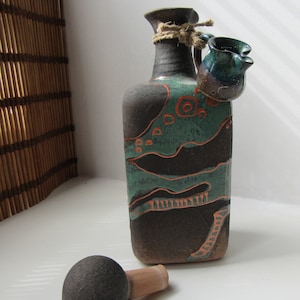 Puede incluir: Una botella de cerámica artesanal con un tapón a juego. La botella es de color marrón oscuro con detalles en verde azulado y naranja, y una pequeña jarra adjunta. El tapón es gris oscuro con un corcho marrón claro.