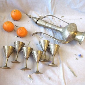 May include: An antique brass pitcher with ornate detailing and six matching goblets. Three orange tangerines are arranged near the pitcher and goblets. The pitcher has a long spout and a handle. The goblets have a floral design.